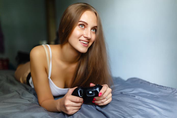 Girl gamer plays with a wireless gamepad while looking at the screen in front of her. Young blonde girl smiles and enjoys winning with gamepad in hand, playing video games console, lying on the bed.